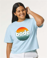 Smiling woman wears designerapp’s BODE Summer Camp 2026 "OG Logo" Women’s Crop Top (Comfort Colors 3023CL) in light blue, featuring a sunset graphic.