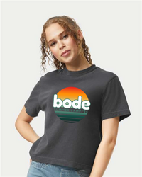 A person wearing the designerapp BODE Summer Camp 2026 "OG Logo" women's black crop top, featuring a sunset graphic and "bode SUMMER CAMP '26" on the front.