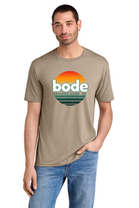 A man wears the Printify 2026 Bode Summer Bootcamp OG Logo Unisex T-Shirt DM130 in beige, featuring a sunset graphic and "bode Summer Camp '23" text. The tri-blend fabric is lightweight, breathable, and perfect for casual summer style with jeans.