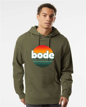A person wears the 2026 Bode Summer Bootcamp — OG Logo Hoodie Sweatshirt by Printify, an olive green unisex fleece hoodie with sunset graphic and logo on front, featuring an adjustable hood, made from Independent Trading Co. SS4500.