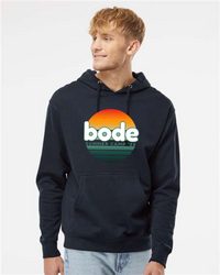 A person in a black Printify 2026 Bode Summer Bootcamp — OG Logo Hoodie Sweatshirt (Unisex Independent Trading Co. SS4500) with a sunset graphic stands looking to the side.
