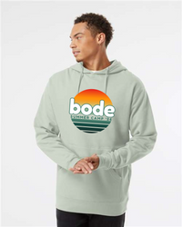 A person stands against a plain background wearing the Printify 2026 Bode Summer Bootcamp OG Logo Hoodie Sweatshirt (Unisex Independent Trading Co. SS4500) in light green, featuring a front sunset graphic and "bode summer camp '26" logo.