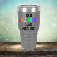 A stainless steel travel mug with a clear lid is displayed on a wooden surface against a blurred background. The Custom UV Printed Drinkware - Single Side Printing Included in Price by Kodiak Coolers features text that reads, "Your full color logo goes here.