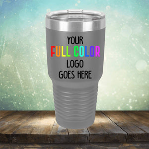 A stainless steel travel mug with a clear lid is displayed on a wooden surface against a blurred background. The Custom UV Printed Drinkware - Single Side Printing Included in Price by Kodiak Coolers features text that reads, "Your full color logo goes here.