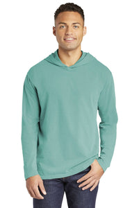 A man stands facing forward, wearing a Comfort Colors Ring Spun Long Sleeve Hoodie Shirt 4900 in light teal and dark jeans, with one hand in his pocket.