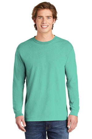 A smiling young man in a Comfort Colors Heavyweight Ring Spun Long Sleeve T-Shirt 6014 light teal and blue jeans stands against a plain white background.