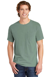 A smiling young man wears a sage green Comfort Colors Heavyweight Ring Spun Pocket T-Shirt 6030 and blue jeans, standing with one hand in his pocket.