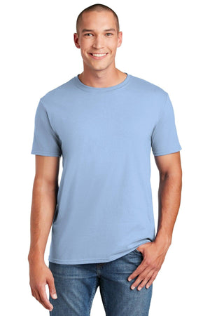 A person wears a Gildan Softstyle T-Shirt 64000 in plain light blue, made from ringspun cotton, with blue jeans, smiling against a white backdrop.