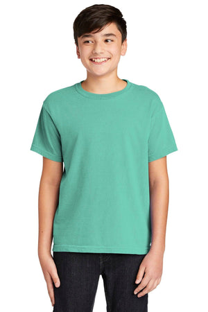 A teen boy in a vintage look Comfort Colors Youth Ring Spun T-Shirt 9018 in green and black pants smiles with his hands by his sides.