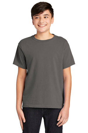 A smiling boy with short dark hair wears the Comfort Colors Youth Ring Spun T-Shirt 9018 in plain dark gray, paired with dark pants, against a white background. The t-shirt is made of 100% ring spun cotton by Comfort Colors.
