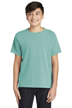 A teen in a Comfort Colors Youth Ring Spun T-Shirt 9018 with a vintage teal look and dark pants smiles against a plain white background.