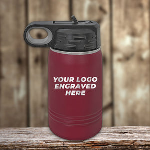 A maroon 12 oz Kodiak Coolers Custom Kids Water Bottle with a black lid, personalized with "YOUR LOGO ENGRAVED HERE" in white on the front, sits on wood—ideal for Black Friday custom logo bottles.