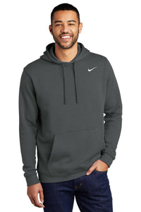 A man wearing a dark green Nike Club Fleece Pullover Hoodie Sweatshirt CJ1611, featuring an embroidered swoosh logo, stands with one hand in his jeans pocket, smiling against a white background.
