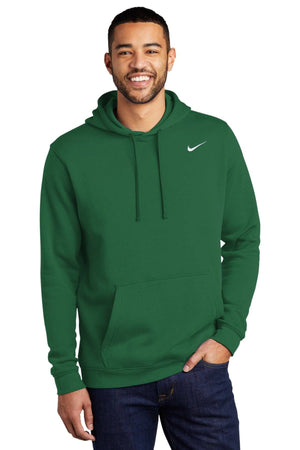 A person wearing a green Nike Club Fleece Pullover Hoodie Sweatshirt CJ1611, featuring an embroidered white Swoosh on the chest, stands against a white background, smiling with one hand in the hoodie pocket.