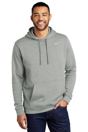A man wearing a light grey Nike Club Fleece Pullover Hoodie Sweatshirt CJ1611 with an embroidered Swoosh smiles, standing with one hand in his pocket against a white background.