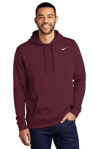 A man wearing a maroon Nike Club Fleece Pullover Hoodie Sweatshirt CJ1611 with an embroidered Swoosh stands against a white background, smiling and placing one hand in the hoodie pocket.