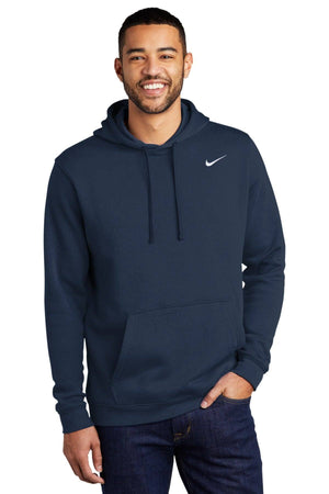A man wearing a dark blue Nike Club Fleece Pullover Hoodie Sweatshirt CJ1611 with an embroidered Swoosh on the front pocket and dark pants smiles at the camera.