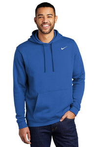 A person wearing a Nike Club Fleece Pullover Hoodie Sweatshirt CJ1611 in blue, featuring an embroidered Swoosh and front pocket, paired with dark jeans, is smiling at the camera.