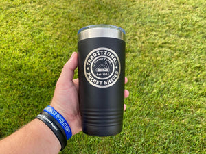 A hand holds a black 20 oz Kodiak Coolers tumbler, custom laser engraved, while wearing blue wristbands. Green grass in the background highlights this set of 6 tumblers—perfect for stylish, low-volume custom orders with free shipping.
