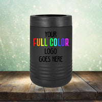 Black cylindrical can cooler with the text "YOUR FULL COLOR LOGO GOES HERE" displayed in colorful letters on the front, set against a bokeh background and wooden tabletop, ideal for pairing with your Kodiak Coolers Custom UV Printed Drinkware - ONE TIME SPECIAL OFFER.