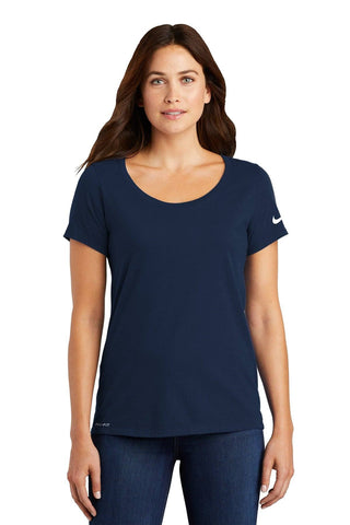 A woman wears a navy Nike Ladies Dri-FIT Cotton/Poly Scoop Neck T-Shirt (NKBQ5234) and jeans, standing against a plain white background.