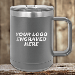The Kodiak Coolers SAMPLE - 15 oz Insulated Coffee Mug, featuring a "YOUR LOGO ENGRAVED HERE" message, is perfect as a promotional gift. The gray metal mug with handle sits on wood against a blurred wooden background.