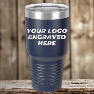 A navy Realtor Closing Gift tumbler by Kodiak Coolers, customized with your company logo engraved on the front and a clear lid, ideal as a promotional drinkware gift, shown on a wooden surface with a blurred wooden background.