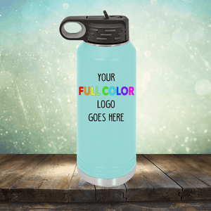 A blue Custom UV Printed Drinkware - ONE TIME SPECIAL OFFER with a black lid by Kodiak Coolers is placed on a wooden surface. The bottle features the text "Your Full Color Logo Goes Here" printed on it, perfect for matching with custom coffee mugs. The background is a blurred, sparkly green scene.
