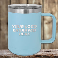 A light blue 15 oz insulated stainless steel coffee mug by Kodiak Coolers sits on a wooden surface, featuring custom laser-engraved text "YOUR LOGO ENGRAVED HERE" in white capital letters. Available with special bulk wholesale pricing.