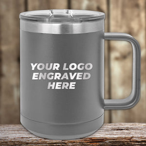 The Kodiak Coolers 15 oz gray stainless steel coffee mug features a laser-engraved "Your Logo Here" text, complete with a clear lid, on a wooden surface. Available with special bulk wholesale pricing.