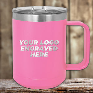 A pink 15 oz Kodiak Coolers insulated mug with a lid, displaying the text "YOUR LOGO ENGRAVED HERE," rests on a wooden table against a blurred background. Crafted from stainless steel, this custom mug prominently features your laser-engraved logo at special bulk wholesale pricing.