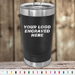 A Kodiak Coolers black insulated tumbler with a laser-engraved "YOUR LOGO ENGRAVED HERE" sits on wood among diverse colors. Ideal for custom orders with no setup fees and attractive wholesale pricing during the Presidents Day sale. Note: $250 minimum order required.