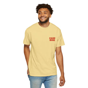 A man wearing a light yellow Printify BODE Summer Camp T-Shirt 2026 "Throwback Sunset" (Unisex Comfort Colors 1717) and blue jeans stands smiling against a plain white background.