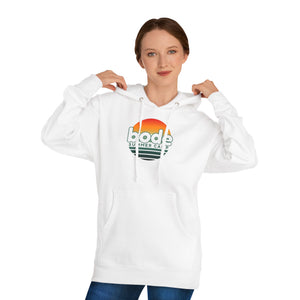 A woman faces forward against a plain background, adjusting the hood of the Printify 2026 Bode Summer Bootcamp — Cali Sunset Hoodie Sweatshirt - SS4500, crafted from soft cotton-poly fleece with a "bode summer camp" logo.