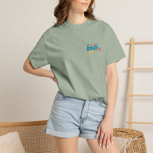 A woman indoors by a wicker chair and wooden rack wears the Printify BODE Summer Camp T-Shirt 2026 — "Life's Better Together" (sage green, Comfort Colors 1717) with light denim shorts.