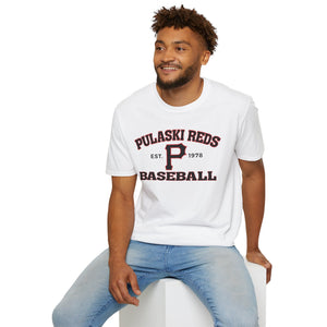 A man in a Printify "P Reds Baseball Est. 1978 - Unisex Softstyle T-Shirt Volume" and jeans sits on a white block, smiling and looking slightly to the side.
