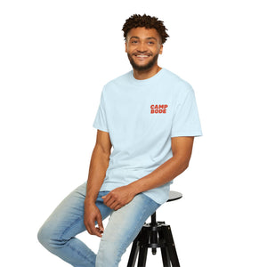 A man in a BODE Summer Camp T-Shirt 2026 "Throwback Sunset" by Printify and jeans sits on a black stool, smiling at the camera against a plain white background.