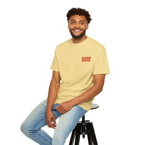 A man wearing a light yellow Printify BODE Summer Camp T-Shirt 2026 "Throwback Sunset" (Unisex Comfort Colors 1717) and light blue jeans sits on a black stool against a white background, smiling at the camera.