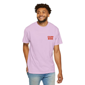 A man smiles against a plain white background, wearing the Printify BODE Summer Camp T-Shirt 2026 "Throwback Sunset" (Unisex Comfort Colors 1717) in light purple with "Camp Bode," paired with blue jeans.