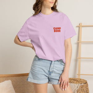 A woman wears the BODE Summer Camp T-Shirt 2026 "Throwback Sunset" by Printify with denim shorts, standing indoors near a wooden rack and wicker furniture.