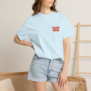 A woman stands indoors with one hand on her hip, wearing a light blue BODE Summer Camp T-Shirt 2026 "Throwback Sunset" by Printify (Unisex Comfort Colors 1717) and denim shorts.