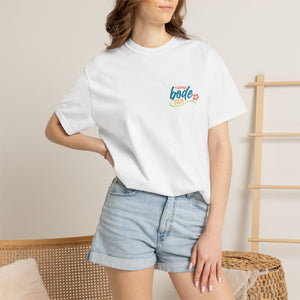 A woman in a white Printify "Summer Camp T-Shirt — 'Life Is Better Together' Beach Can Design" and denim shorts stands by a wooden chair and decorative ladder in a bright, modern room.