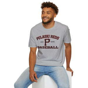 A man wearing a Printify P Reds Baseball Est. 1978 Unisex Softstyle T-Shirt in gray and light blue jeans sits on a white cube, smiling and looking to the side.