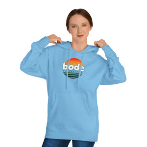 A woman adjusts the hood of a Printify 2026 Bode Summer Bootcamp — Cali Sunset Hoodie Sweatshirt (SS4500) featuring a "bode summer camp" logo and sunrise graphic, standing against a plain white background.