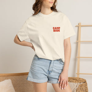 A woman stands indoors beside a wooden rack and wicker chair, wearing the Printify BODE Summer Camp T-Shirt 2026 "Throwback Sunset"—a white Unisex Comfort Colors 1717 heavyweight cotton tee—and light denim shorts.