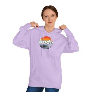 A person faces forward, holding the hood strings of a light purple Printify 2026 Bode Summer Bootcamp — Cali Sunset Hoodie Sweatshirt (SS4500) featuring a “bode summer camp” logo.