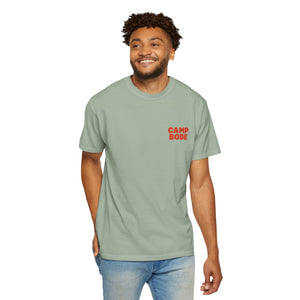 A man stands smiling, wearing the Printify BODE Summer Camp T-Shirt 2026 "Throwback Sunset" (light green, Unisex Comfort Colors 1717) with "CAMP BODE" in red on the chest, and light blue jeans.