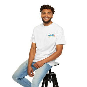 A man wearing the Printify Summer Camp T-Shirt — 'Life Is Better Together' Beach Can Design pairs it with light blue jeans, sitting on a black stool against a white background and smiling at the camera.
