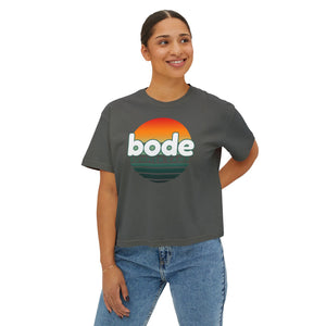 A woman smiles, wearing blue jeans and a Printify 2026 Bode Summer Bootcamp - Cali Sunset Logo Crop T-Shirt (3023CL), featuring a sunset graphic and "bode SUMMER CAMP" text on the front.