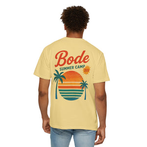 A person wears the Printify BODE Summer Camp T-Shirt 2026 — "Throwback Sunset," a unisex Comfort Colors 1717 tee featuring “Bode Summer Camp 2026” and a sunset, palm trees, and stripes graphic on the back.